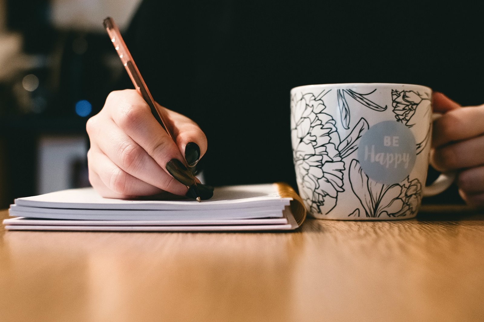 A white person's hand with black nails writes in an open journal with a pen. Next to them is a floral mug.